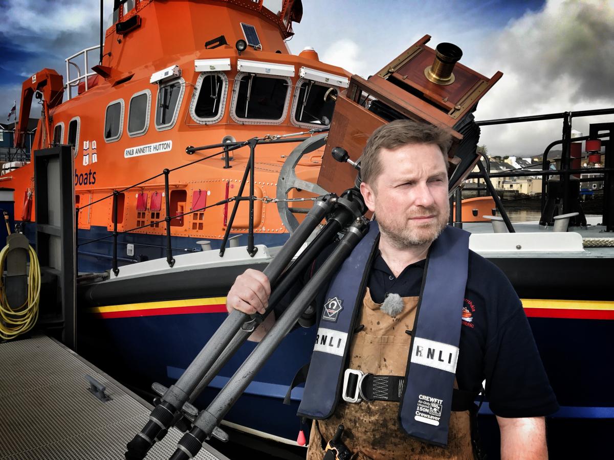 A photographer stands in front of an RNLI orange lifeboat holding an Edwardian camera on a tripod 